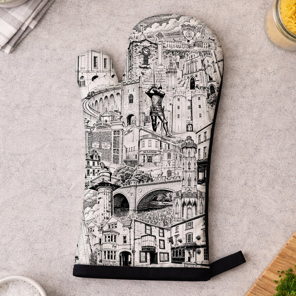A single oven mitt laid flat on a light gray surface, printed with a detailed black-and-white architectural cityscape illustration. Surrounding props include a small bowl of salt, chopped parsley on a cutting board, a folded napkin with a plant leaf, and kitchen items in the corners.