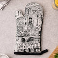 A single oven mitt laid flat on a light gray surface, printed with a detailed black-and-white architectural cityscape illustration. Surrounding props include a small bowl of salt, chopped parsley on a cutting board, a folded napkin with a plant leaf, and kitchen items in the corners.