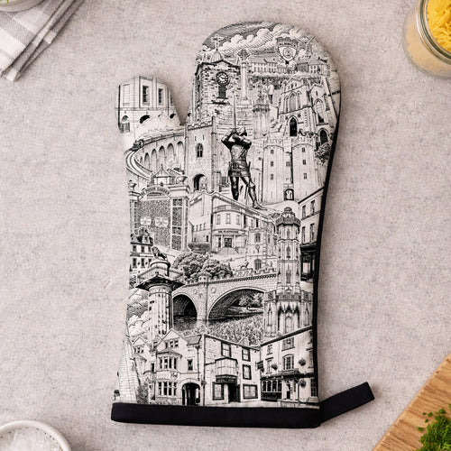 A single oven mitt laid flat on a light gray surface, printed with a detailed black-and-white architectural cityscape illustration. Surrounding props include a small bowl of salt, chopped parsley on a cutting board, a folded napkin with a plant leaf, and kitchen items in the corners.