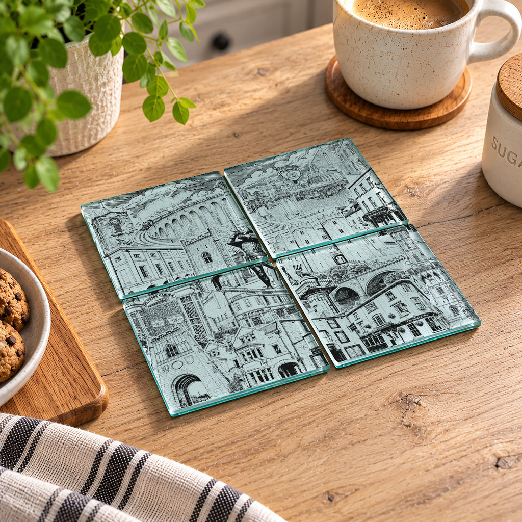 Four square glass coasters with a detailed architectural illustration print, arranged in a 2x2 grid on a wooden table next to a coffee cup, a plant, and a striped cloth.