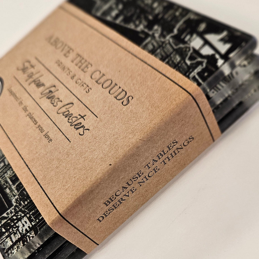 A pack of glass coasters with a white cityscape pattern, wrapped in a brown kraft paper band that reads 'Above the Clouds'.