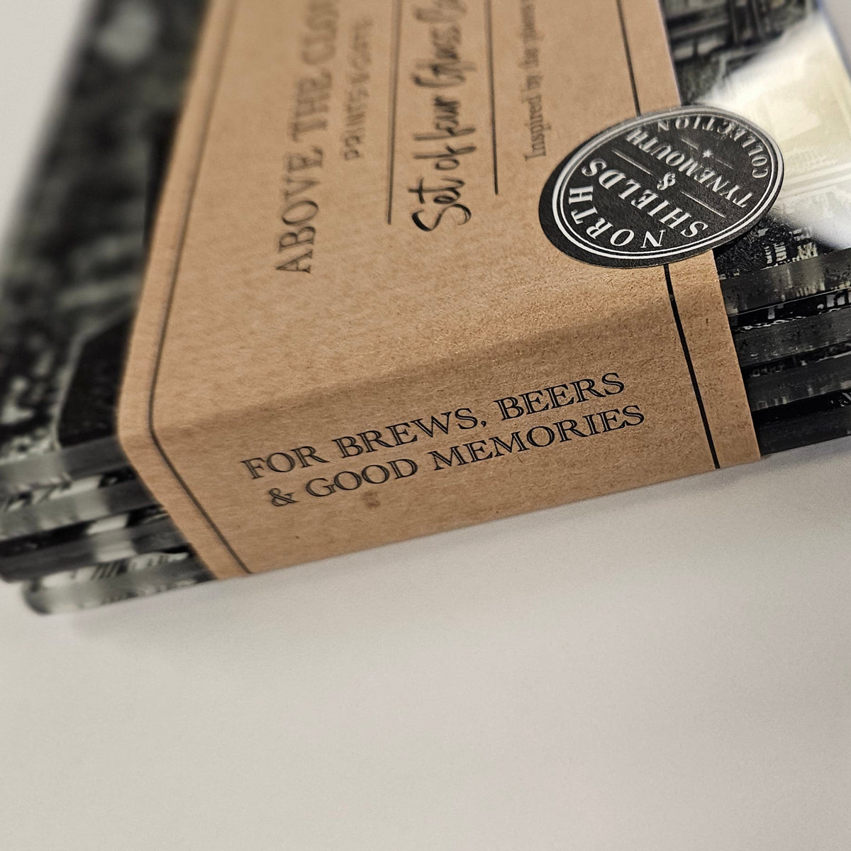 A stack of black textured beer coasters wrapped in a kraft paper band with the brand name 'Above All Things' and the phrase 'For brews, beers & good memories'.
