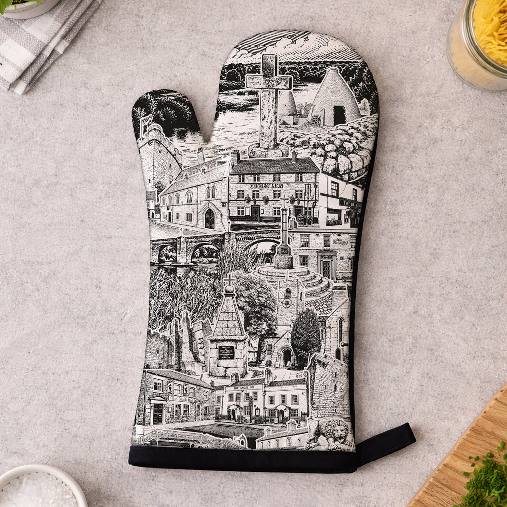 A single oven mitt with a black and white architectural cityscape illustration, laid flat on a light gray countertop with kitchen props around it.