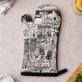 A single oven mitt with a black and white architectural cityscape illustration, laid flat on a light gray countertop with kitchen props around it.
