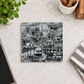 A square coaster featuring a black and white architectural cityscape illustration, displayed on a light marble countertop with kitchen props including a potted plant, a striped cloth, and utensils.
