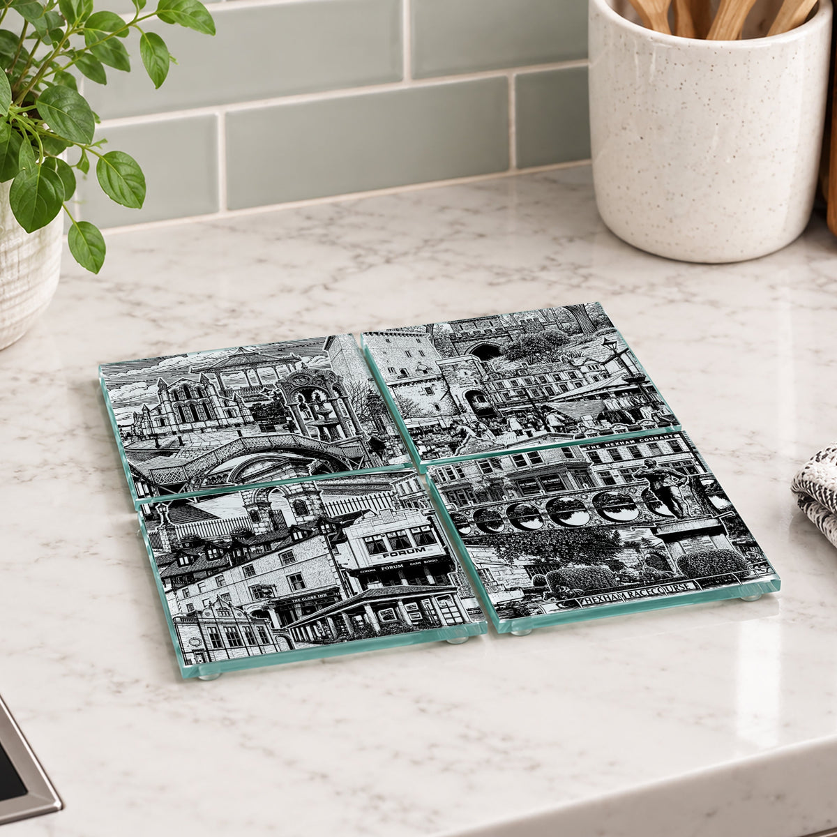 Four square glass coasters with a black and white city street scene print, arranged in a 2x2 grid on a white marble kitchen countertop.