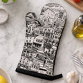 A single oven mitt with a black and white illustrated print of a European city street scene, laid flat on a white marble countertop surrounded by kitchen props including basil, tomatoes, garlic, olive oil, and a striped cloth.