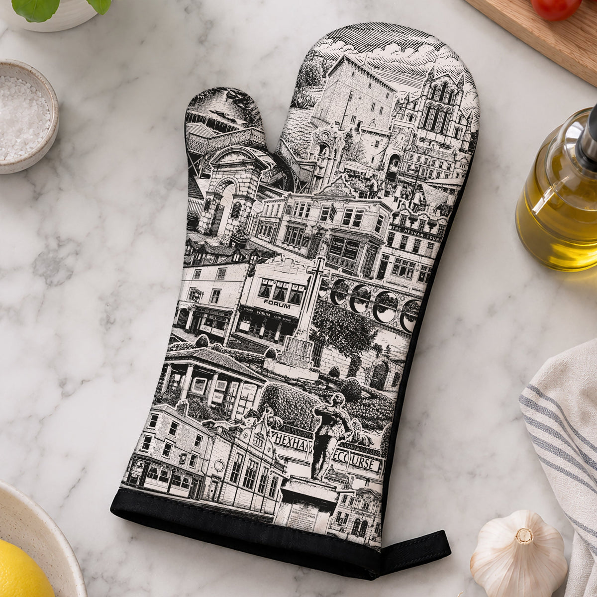A single oven mitt with a black and white illustrated print of a European city street scene, laid flat on a white marble countertop surrounded by kitchen props including basil, tomatoes, garlic, olive oil, and a striped cloth.