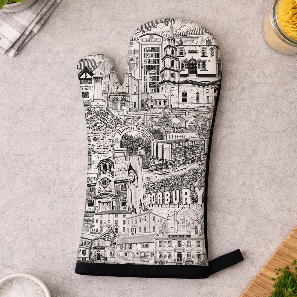 A single oven mitt laid flat on a light gray countertop, printed with a black-and-white illustrated cityscape and the word "HORBURY" near the bottom. Surrounding props include a small bowl of salt, chopped parsley, a wooden cutting board, and kitchen items in the corners.