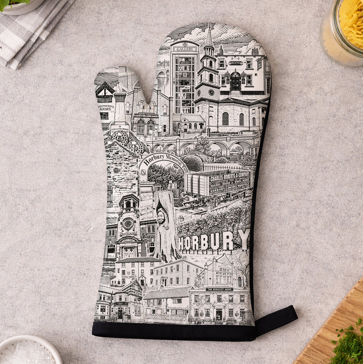 A single oven mitt laid flat on a light gray countertop, printed with a black-and-white illustrated cityscape and the word "HORBURY" near the bottom. Surrounding props include a small bowl of salt, chopped parsley, a wooden cutting board, and kitchen items in the corners.