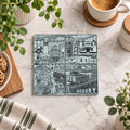 A square ceramic tile coaster featuring a detailed black and white illustration of Horbury local history, displayed on a white marble surface surrounded by lifestyle props like a coffee cup, cookies, and green plants.