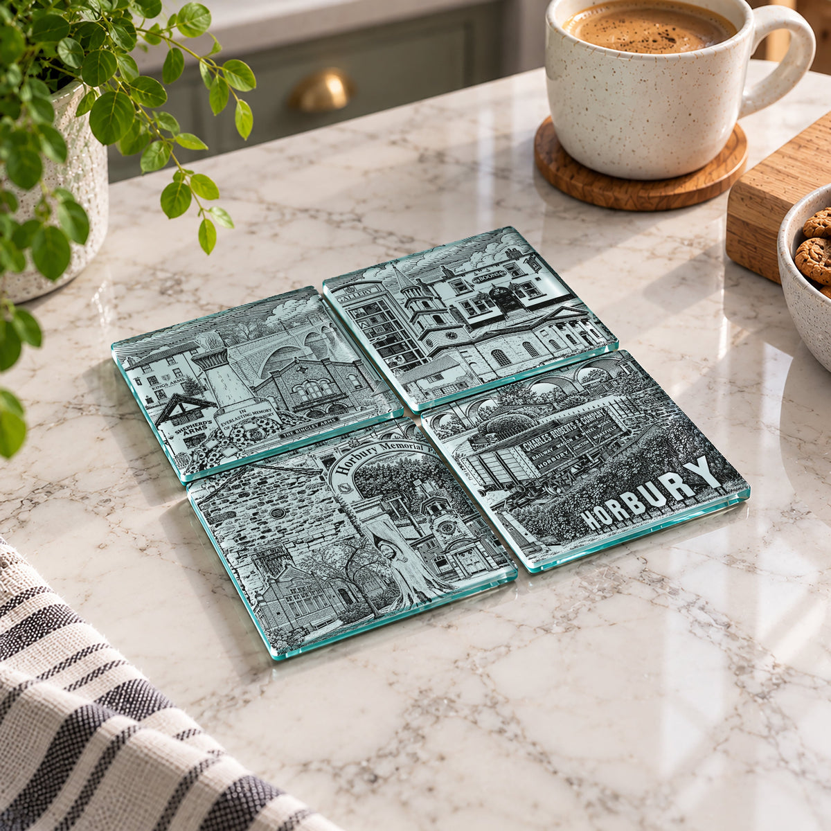 Four square glass coasters with detailed black-and-white architectural illustrations and the word "HORBURY" on one piece, arranged on a white marble countertop with a mug, plant, and kitchen items nearby.