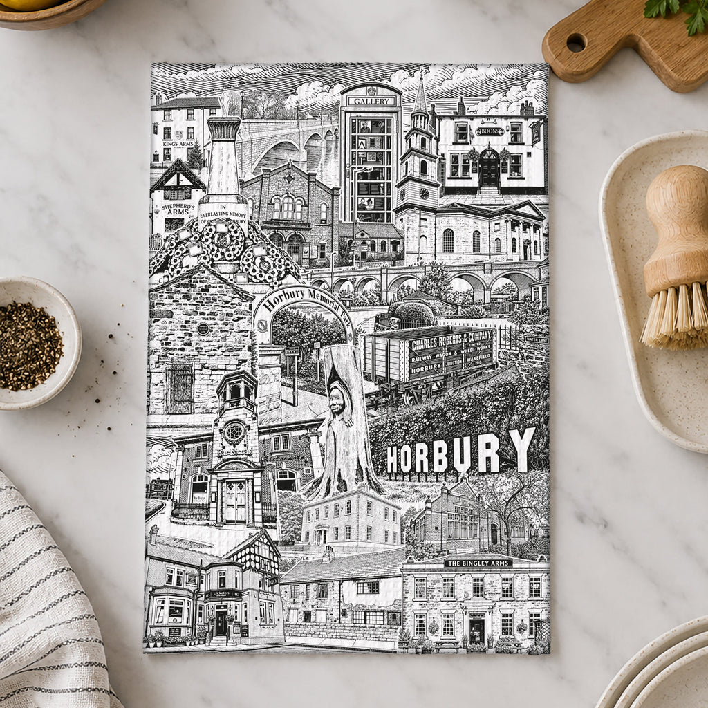 A rectangular black-and-white illustrated print or poster featuring a detailed town scene labeled 'HORBURY,' placed on a light marble surface with kitchen props around it, including lemons, a cream pitcher, herbs, a brush, and stacked plates.