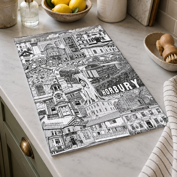 A rectangular kitchen towel featuring a detailed black and white hand-drawn illustration of Horbury, South Yorkshire, styled on a light marble countertop in a bright kitchen setting.