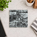 A square ceramic coaster with a detailed black and white industrial collage print, displayed on a light marble kitchen countertop next to a potted plant, a striped cloth, and a stove.