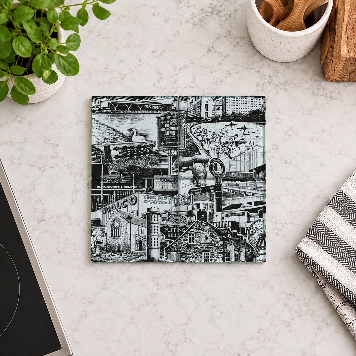 A square ceramic coaster with a detailed black and white industrial collage print, displayed on a light marble kitchen countertop next to a potted plant, a striped cloth, and a stove.