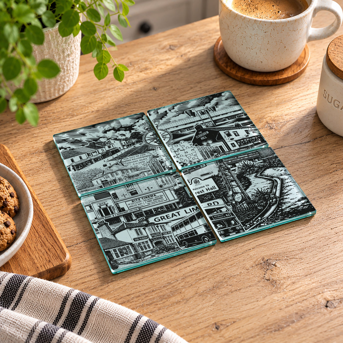 Four square glass coasters with black-and-white illustrated street-scene designs arranged on a wooden table in a real kitchen setting, with a mug, plant, and food visible around them.