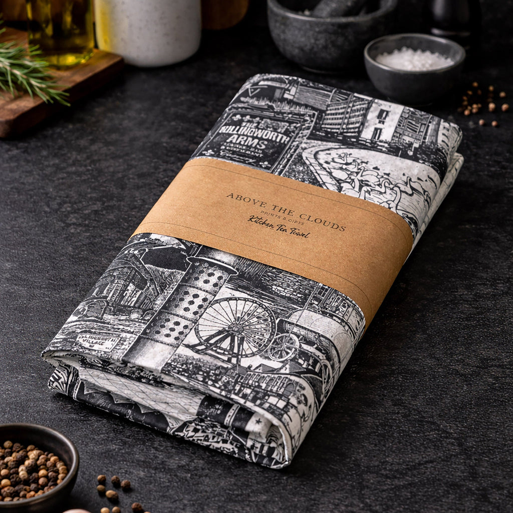 Folded tea towel with black and white design and brown label on a dark surface with kitchen items.