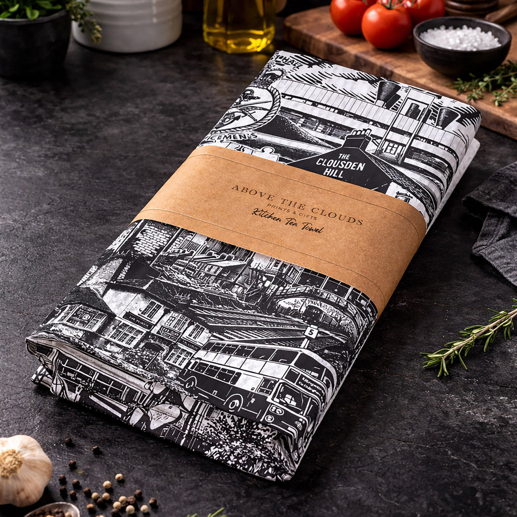 Folded tea towel with black and white pattern and brown label on a dark surface with kitchen items.