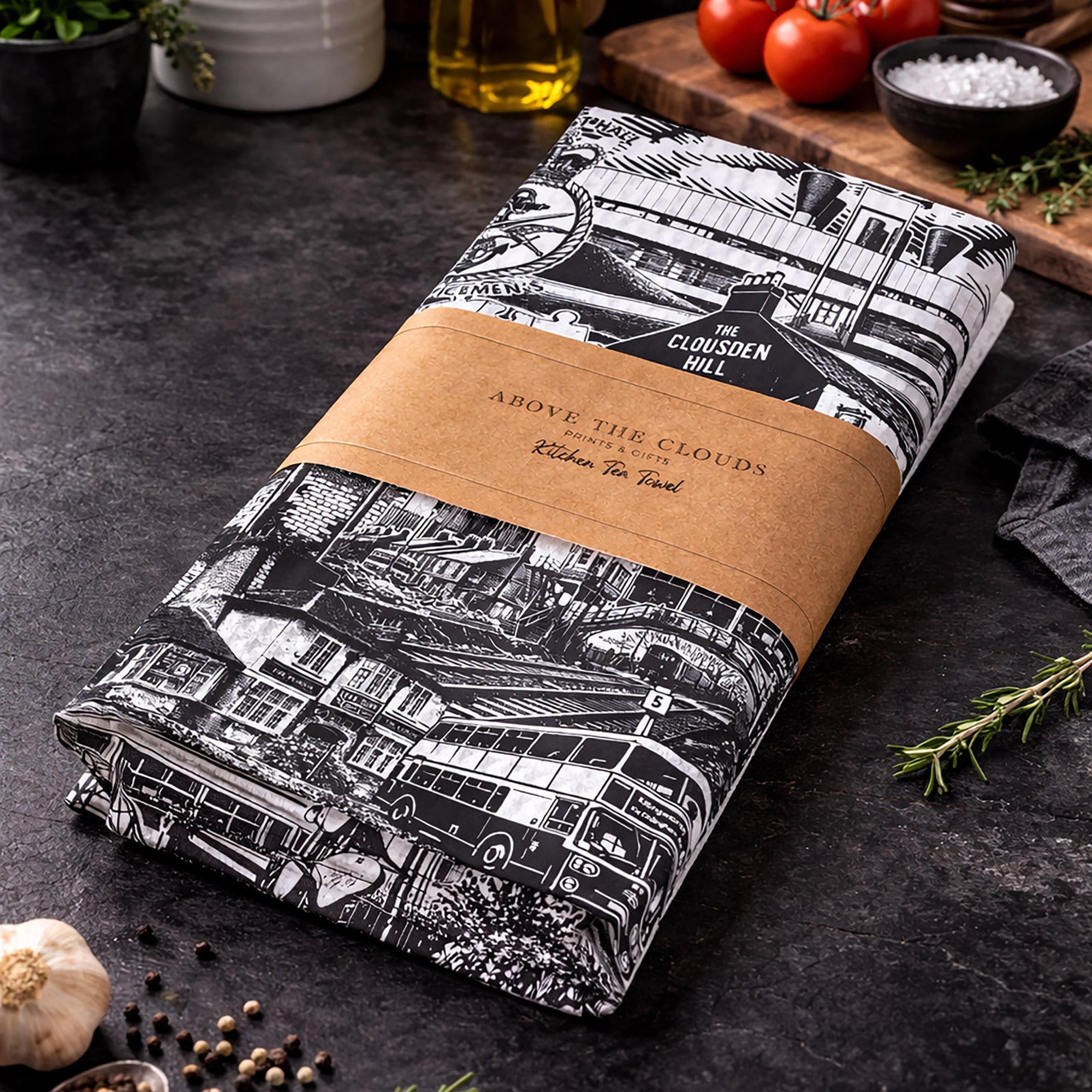 Folded tea towel with black and white pattern and brown label on a dark surface with kitchen items.
