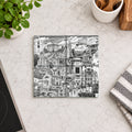 A square black and white illustrated coaster featuring a detailed collage-style drawing of architectural landmarks and figures, displayed on a light marble countertop with kitchen props.