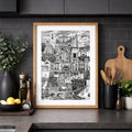 A framed black and white architectural art print of a hillside village, styled on a dark kitchen countertop with kitchen accessories.
