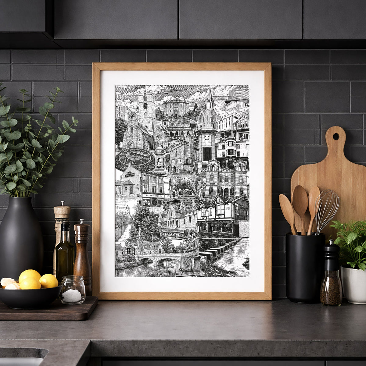 A framed black and white architectural art print of a hillside village, styled on a dark kitchen countertop with kitchen accessories.