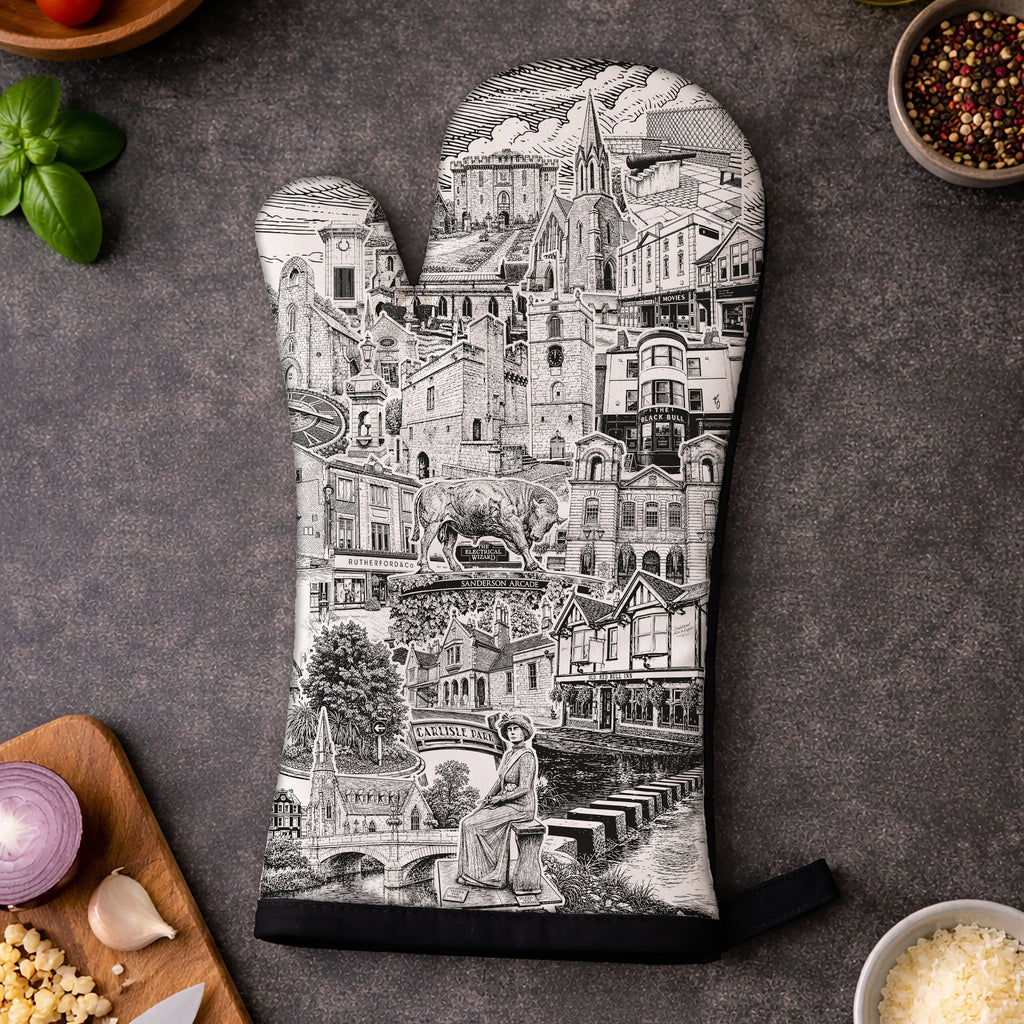 A single oven mitt with a black and white architectural cityscape print is laid flat on a dark gray surface, surrounded by cooking ingredients including basil, tomatoes, garlic, onion, cheese, and a knife.