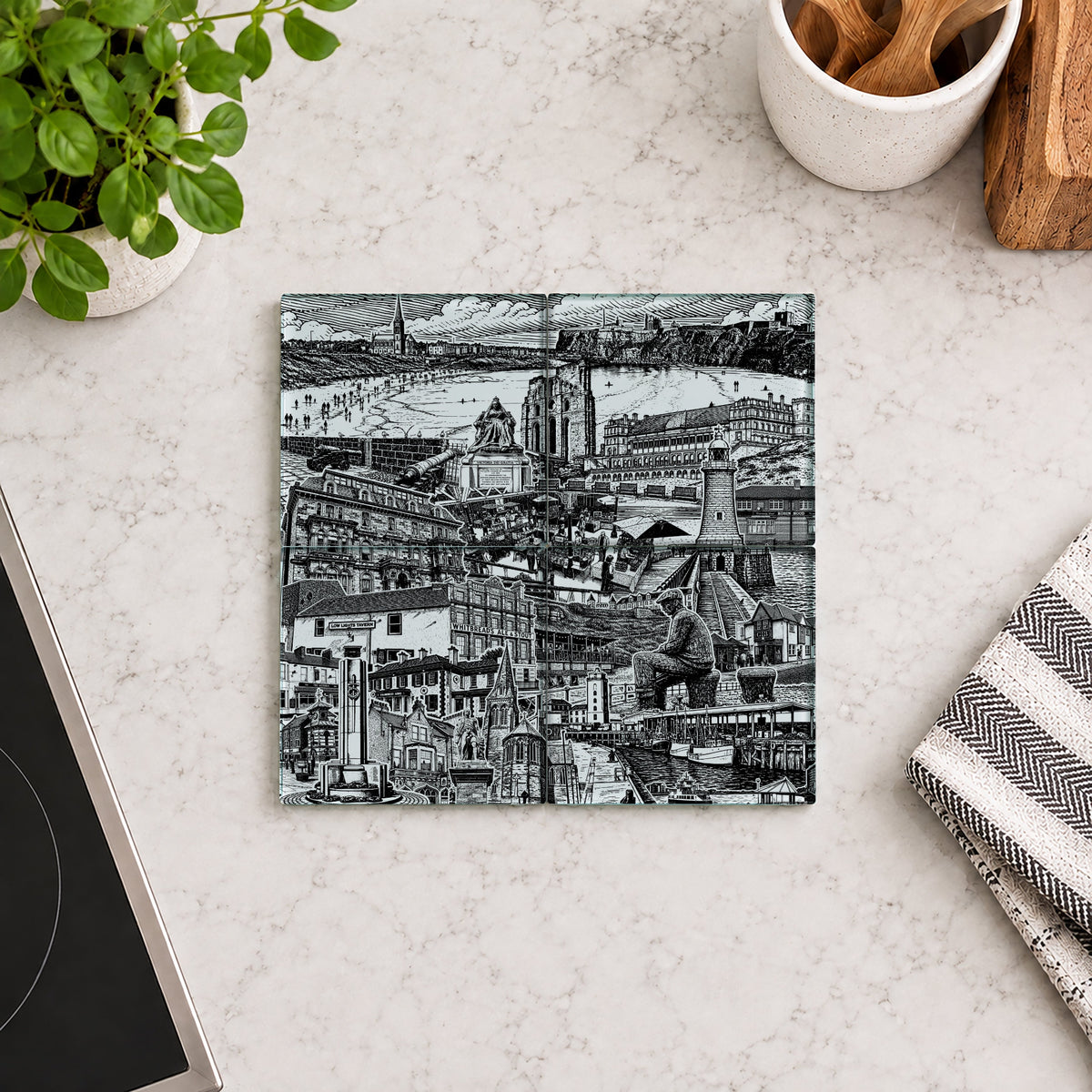 A square black and white illustrated coaster or tile with a detailed cityscape print, placed on a light marble countertop with kitchen props including a potted plant, utensils in a cup, and a striped cloth.