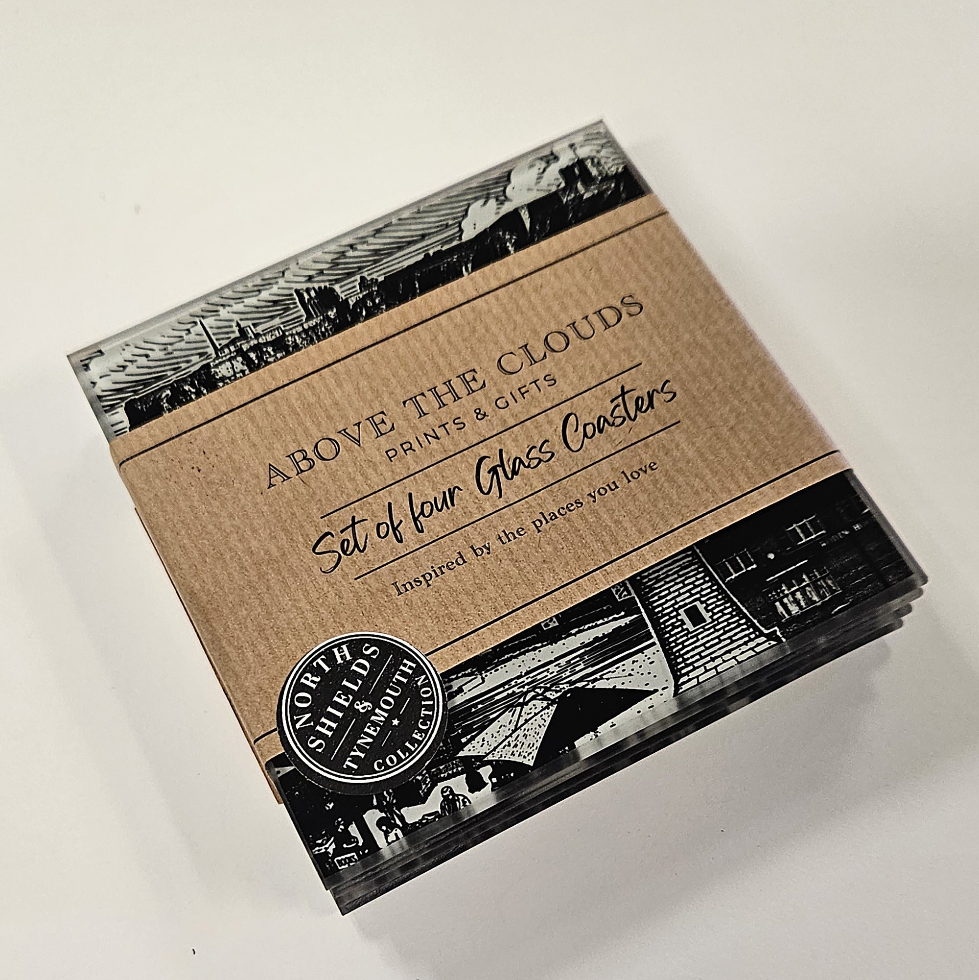 A boxed set of four glass coasters by Above the Clouds Prints & Gifts, shown on a plain light background. The packaging is kraft paper with black-and-white architectural illustrations and a round black-and-white logo seal.