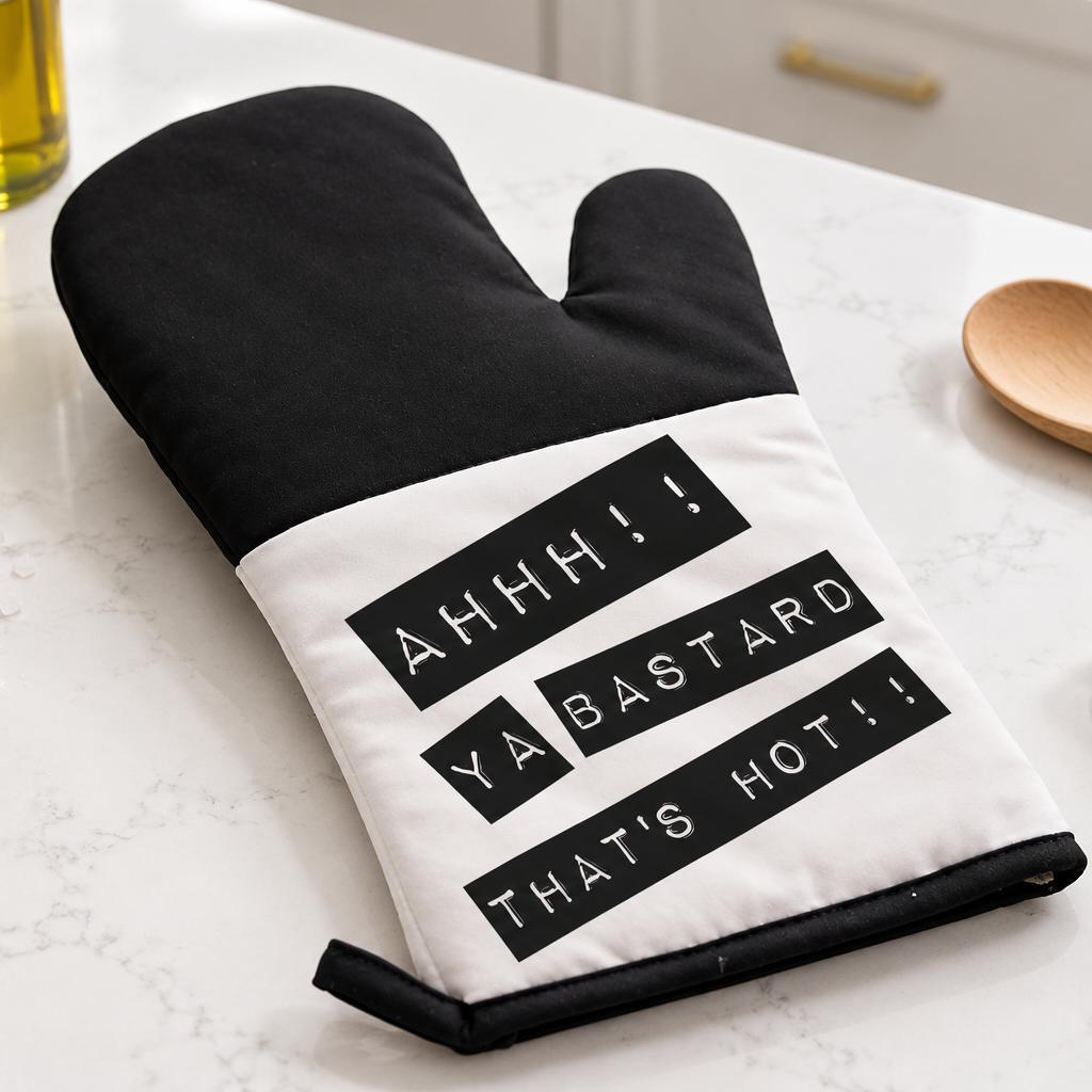 A black and white oven mitt with the text 'AHHH!! YA BASTARD THAT'S HOT!!' printed on it, styled on a white marble countertop with kitchen props like a wooden spoon, green towel, and basil plant.