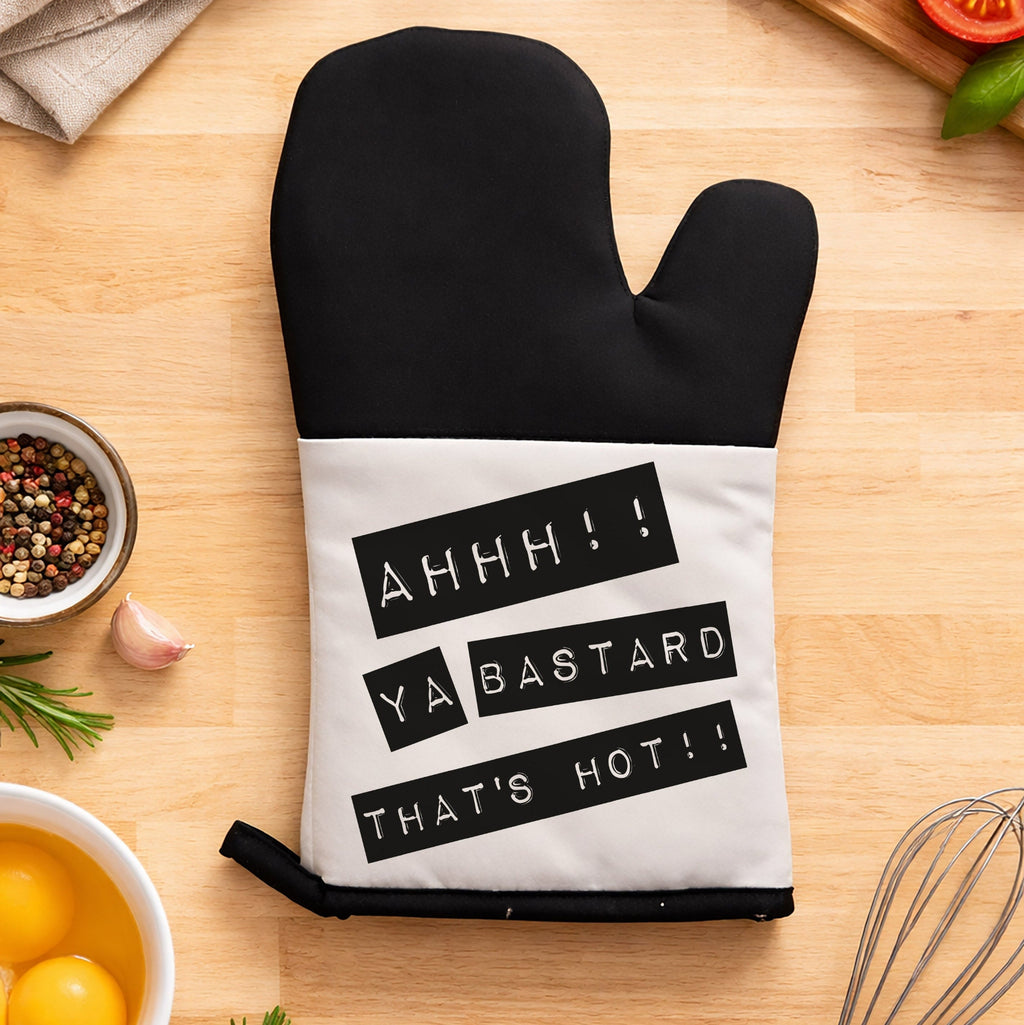 Black oven mitt with humorous text on a kitchen counter with cooking ingredients.