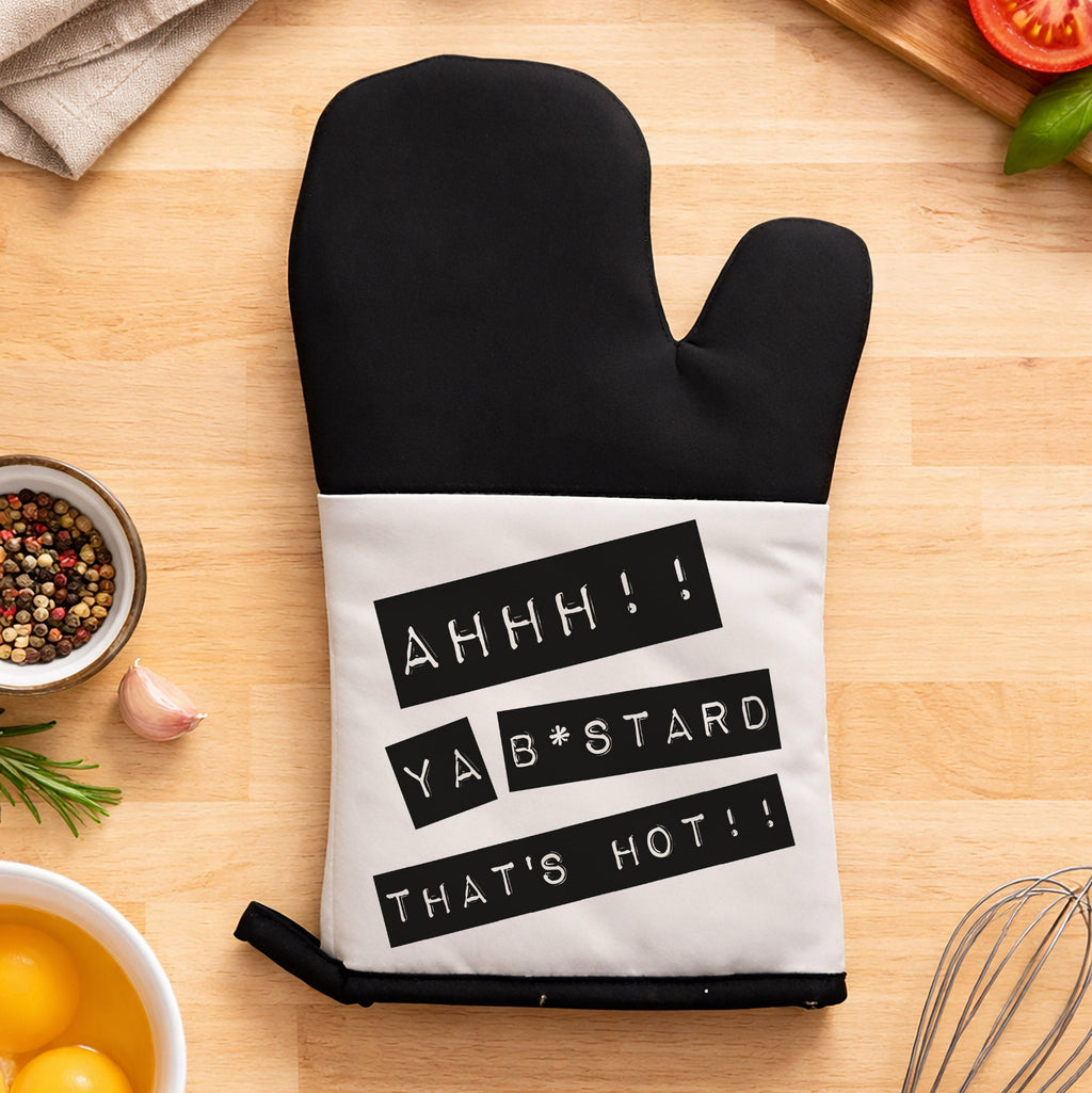 Black oven mitt with humorous text on a kitchen counter with cooking ingredients.