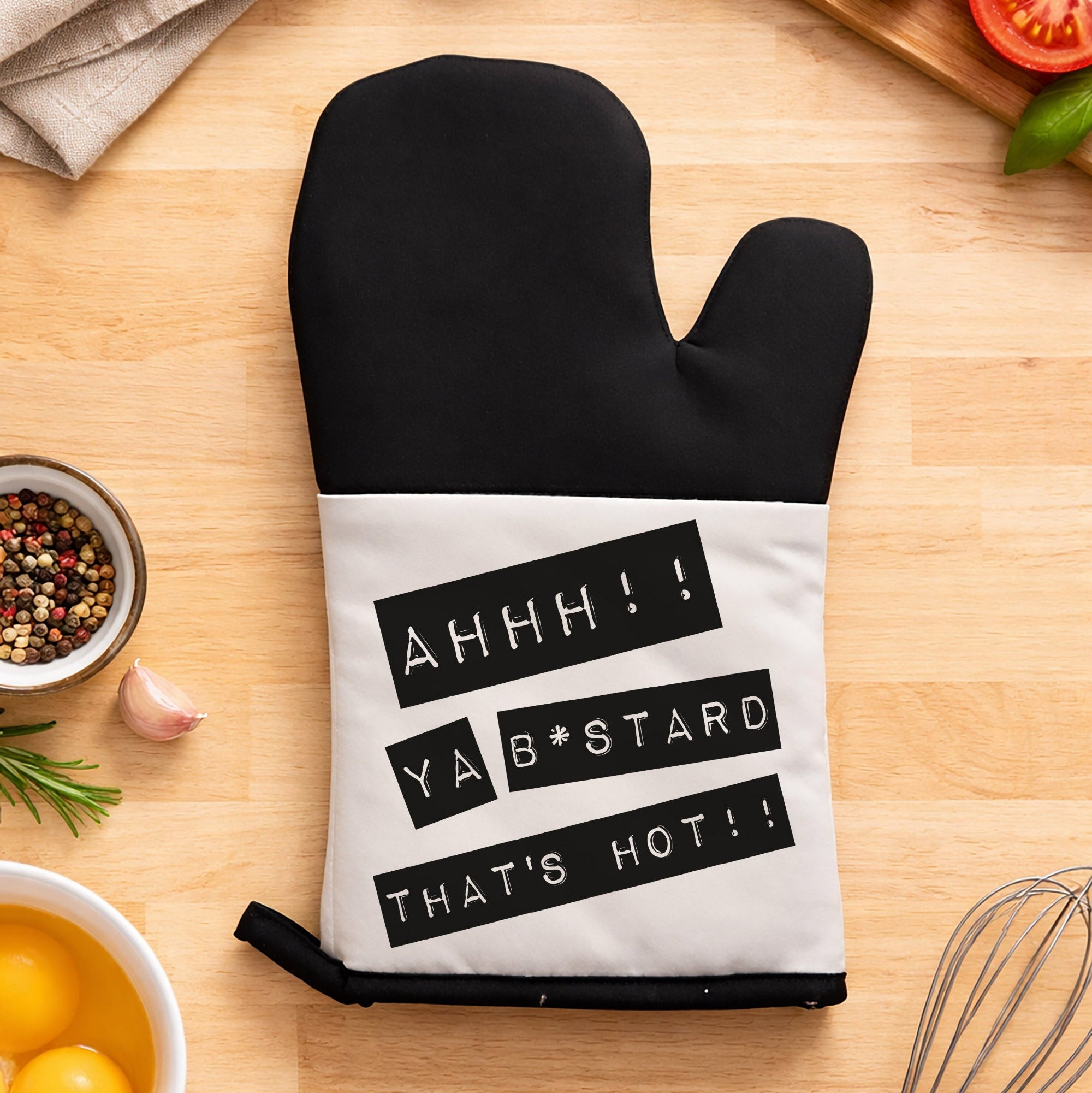 Black oven mitt with humorous text on a kitchen counter with cooking ingredients.