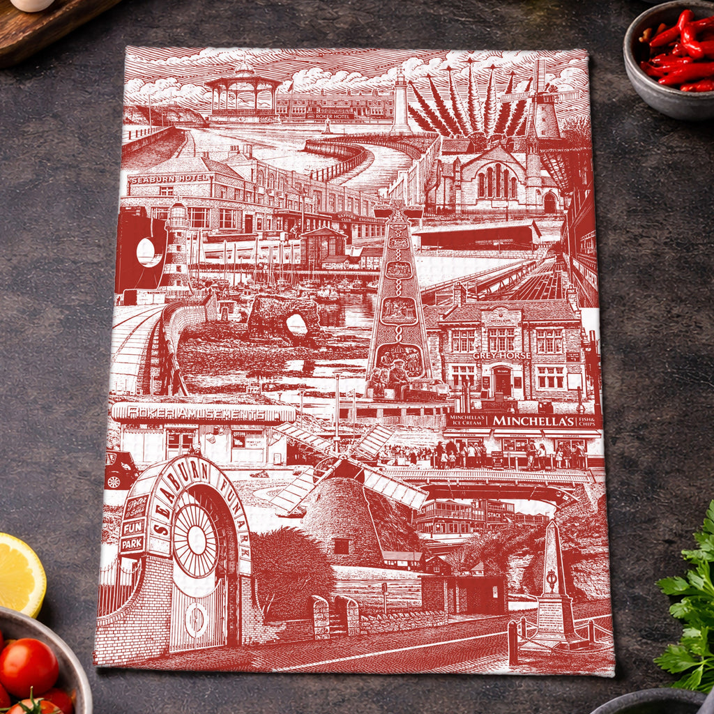 A rectangular kitchen towel with a red and white Sunderland themed architectural collage print, styled on a dark textured surface with food props like tomatoes, basil, garlic, olives, chili peppers, and a lemon.