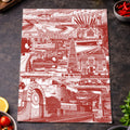 A rectangular kitchen towel with a red and white Sunderland themed architectural collage print, styled on a dark textured surface with food props like tomatoes, basil, garlic, olives, chili peppers, and a lemon.