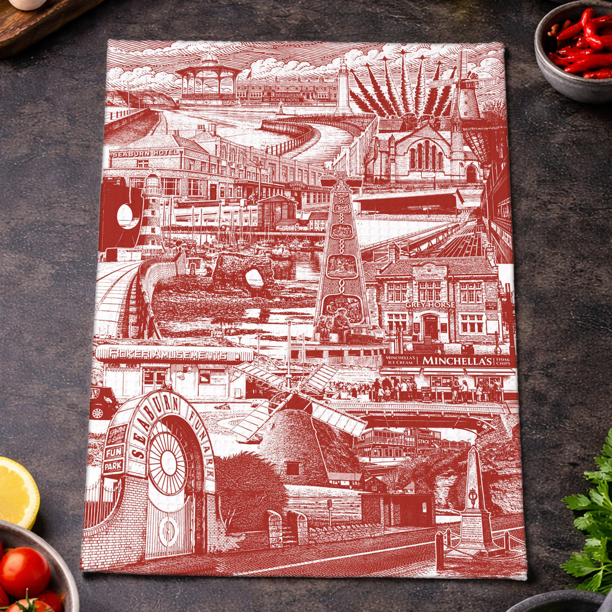 A rectangular kitchen towel with a red and white Sunderland themed architectural collage print, styled on a dark textured surface with food props like tomatoes, basil, garlic, olives, chili peppers, and a lemon.