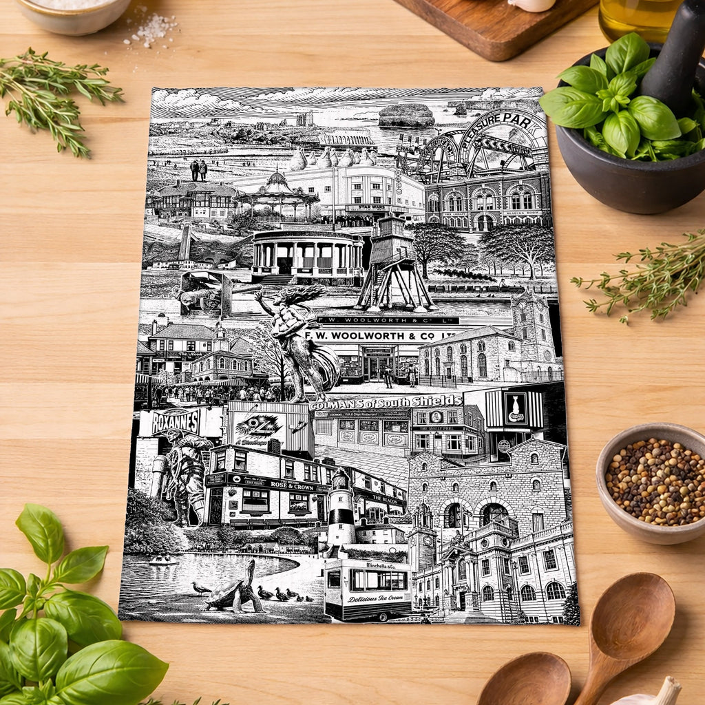 A rectangular black-and-white illustrated tea towel laid on a wooden tabletop, surrounded by kitchen ingredients and utensils including basil, tomatoes, garlic, olive oil, spices, wooden spoons, and a mortar and pestle. 