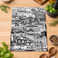 A rectangular black-and-white illustrated tea towel laid on a wooden tabletop, surrounded by kitchen ingredients and utensils including basil, tomatoes, garlic, olive oil, spices, wooden spoons, and a mortar and pestle. 