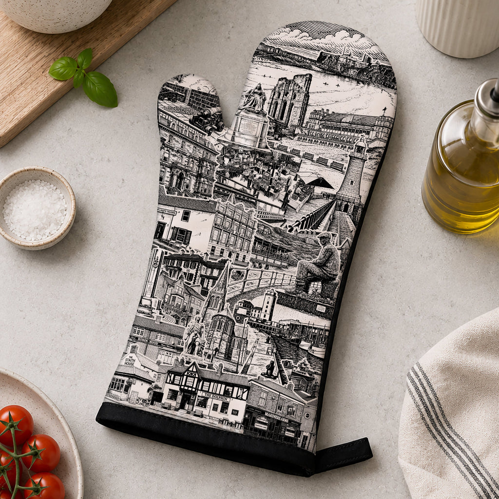 A single oven mitt with a black and white illustrated cityscape print is laid flat on a light stone surface, surrounded by kitchen props including a basil plant, tomatoes, salt, wooden spoon holder, olive oil bottle, and a striped cloth.