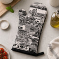 A single oven mitt with a black and white illustrated cityscape print is laid flat on a light stone surface, surrounded by kitchen props including a basil plant, tomatoes, salt, wooden spoon holder, olive oil bottle, and a striped cloth.
