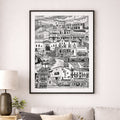 A framed black and white architectural art print of a hillside village, displayed on a light gray wall above a modern beige sofa with decorative pillows in a styled living room setting.