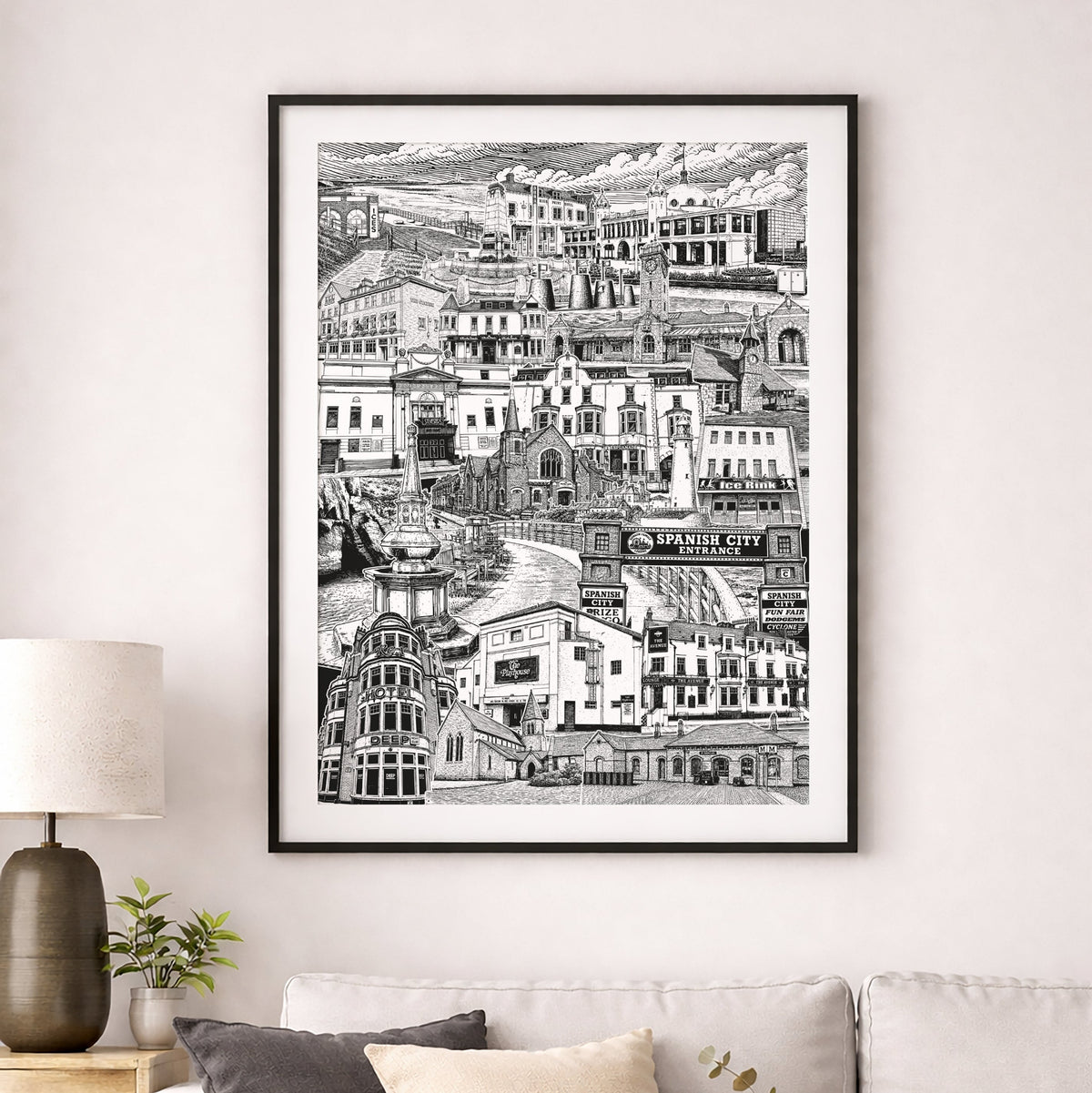 A framed black and white architectural art print of a hillside village, displayed on a light gray wall above a modern beige sofa with decorative pillows in a styled living room setting.