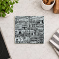 A square ceramic coaster featuring a black and white architectural cityscape illustration, displayed on a light marble countertop with kitchen props including a potted plant, a striped cloth, and a wooden spoon holder.