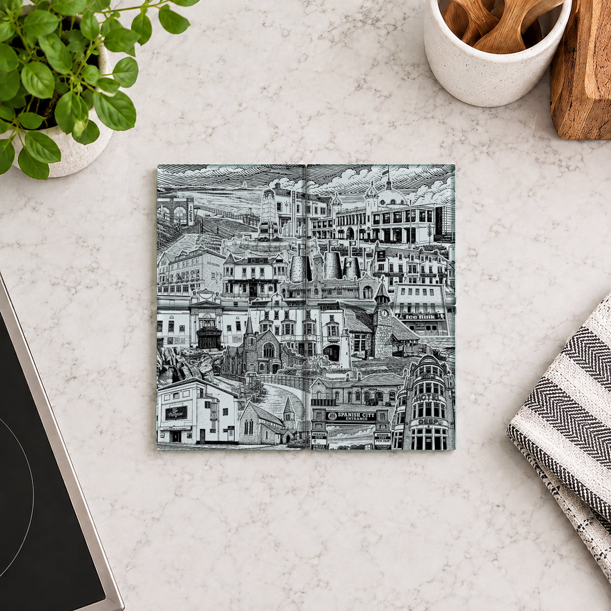 A square ceramic coaster featuring a black and white architectural cityscape illustration, displayed on a light marble countertop with kitchen props including a potted plant, a striped cloth, and a wooden spoon holder.