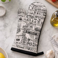 A single oven mitt with a black and white hand-drawn illustration of Venice, Italy, is laid flat on a white marble countertop surrounded by kitchen props like garlic, tomatoes, basil, and olive oil.