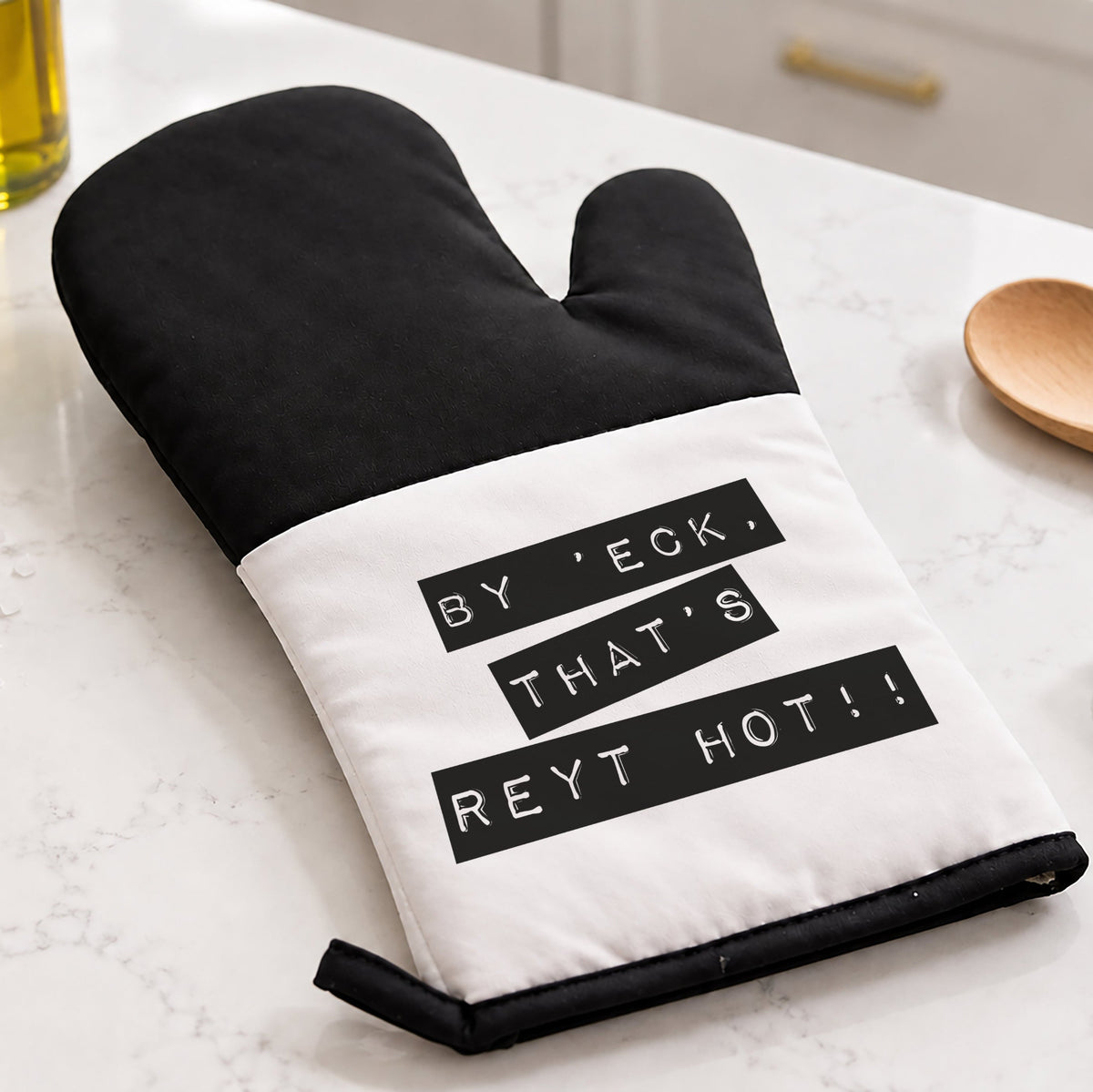A black and white oven mitt with the text 'BY 'ECK, THAT'S REYT HOT!!' printed on the front, resting on a white marble kitchen countertop with a wooden spoon, green towel, and potted plant nearby.